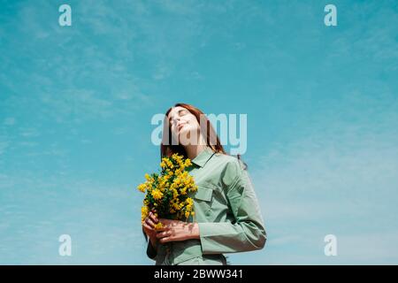 Retrato de una joven roja con los ojos cerrados de pie contra el cielo sosteniendo un montón de flores amarillas
