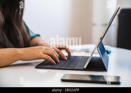 Primer plano de las manos de una mujer escribiendo en el teclado de su tablet híbrido y el dispositivo portátil junto a un teléfono en una mesa, mientras teletrabajo y adaptación t Foto de stock