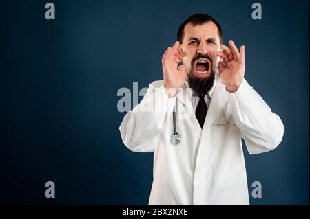 Retrato de un médico masculino con estetoscopio en uniforme médico gritando con las manos hacia la boca posando sobre un fondo azul aislado. Foto de stock