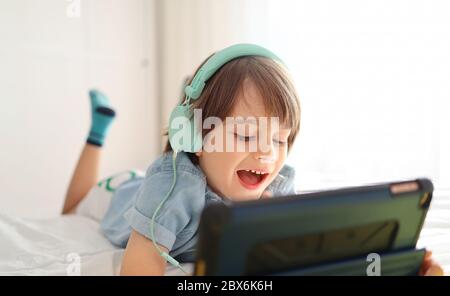 El niño moderno en auriculares está usando una tableta digital y sonriendo mientras se acostaba en su cama en casa - el niño del cute en una camisa azul jugando juegos y watc