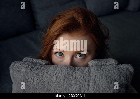Una chica de pelo rojo con ojos azules se asoma por detrás de la almohada. Foto horizontal Foto de stock