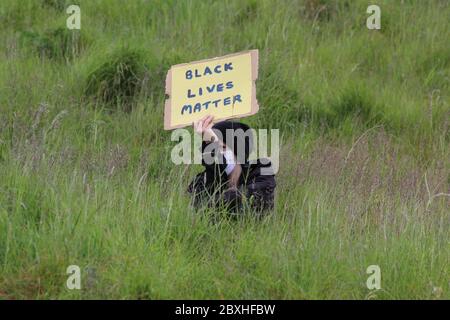Un hombre con una máscara de cara sentado en hierba alta y sosteniendo un cartel leyendo vidas negras importa en una demostración en Edimburgo