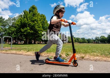 Un niño juega con y empuja su scooter en un día soleado en el parque. Cielo azul y árboles verdes. Niño activo y energético. Foto de stock