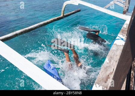 Los buceadores que hacen atrás se voltean al mar azul transparente desde el tablero de un barco. Hay varias maneras de bajar del barco cuando se bucea. Uno de ellos ya está