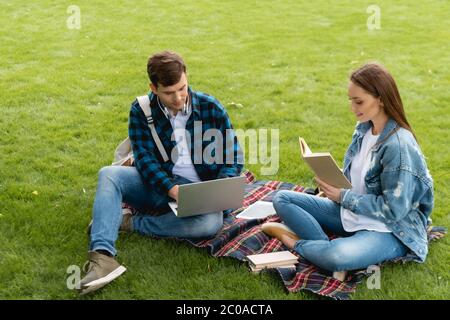 alegre niña leyendo libro cerca de estudiante guapo usando portátil, concepto de estudio en línea Foto de stock