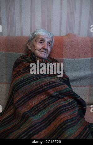 Una anciana está sentada en el sofá envuelto en una manta. Foto de stock