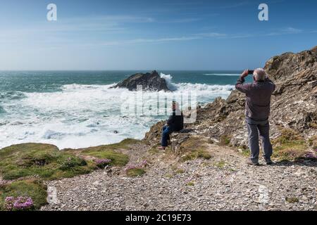Turistas disfrutando del tiempo salvaje y ventoso utilizando un smartphone para fotografiar las olas rompiendo sobre Goose Rock en la costa de Pentire Point East en NE Foto de stock