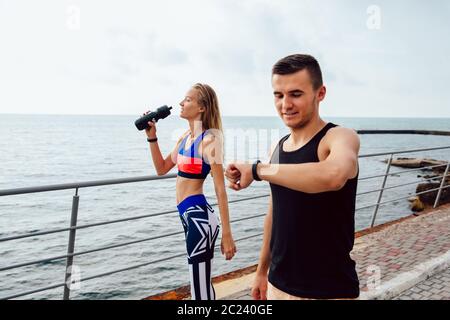 Tío musculoso feliz comprobando un cardio y tiempo después de la carrera, mientras que una niña fitness beber un agua después de ejecutar una distace. En el exterior, cerca del océano. Foto de stock