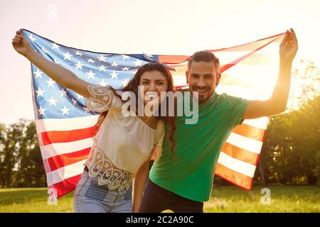 Pareja con la bandera estadounidense celebrando el día de la independencia de américa de pie en el parque. Foto de stock