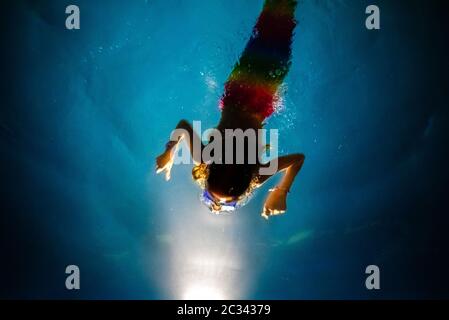 Niña disfrazada de sirena buceando hacia la luz de una piscina azul, con un fondo de ensueño de aguas cristalinas.