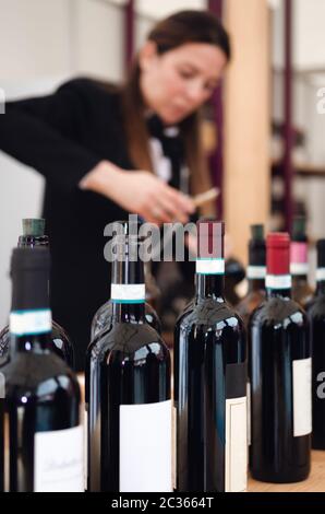 Experiencia de degustación con botellas y una sommelier femenina desenfocada descorpando una botella de vino tinto dolcetto en el fondo Foto de stock