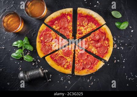 Pizza de pepperoni sobre fondo negro, con copas de vino, albahaca, sal y pimienta, rodada desde arriba Foto de stock