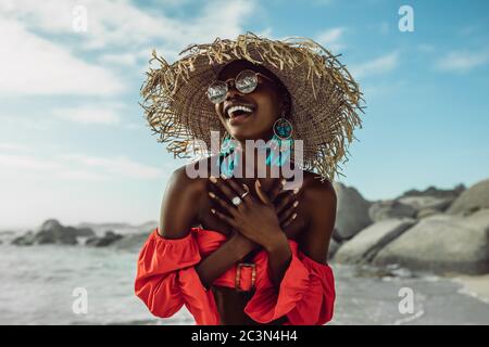 Hermosa mujer africana en vestido rojo con sombrero de paja sonriendo en la playa. Mujer en la sundress disfrutando en la playa.