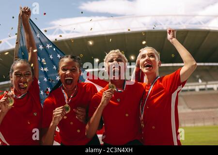 Equipo de fútbol americano femenino celebrando el campeonato. Las mujeres del equipo de fútbol gritando en el campo sosteniendo bandera nacional con confeti caen alrededor.