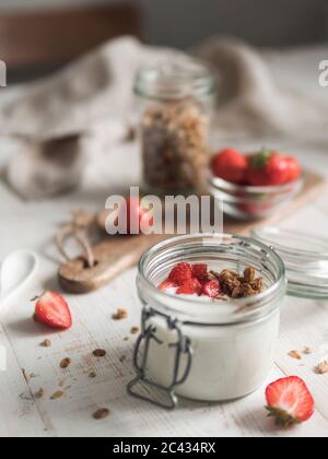 Kéfir probiótico fermentado o yogur en tarro de vidrio servido granola y fresas frescas en mesa rústica de madera blanca. Foto de stock