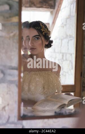 Mujer joven en vestido de boda con libro mirando al espejo