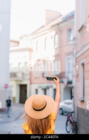Vista trasera de la mujer toma selfie con teléfono inteligente mientras está parado en el callejón en la ciudad Foto de stock