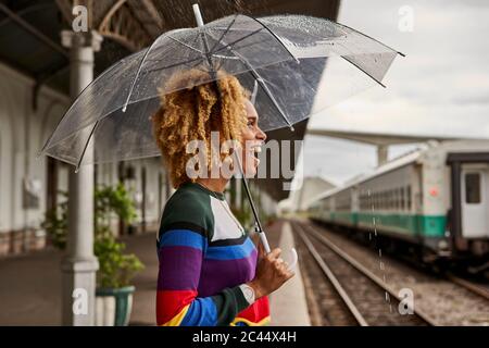 Vista lateral de alegre joven disfrutando mientras se paraba con paraguas en la estación de ferrocarril durante el monzón
