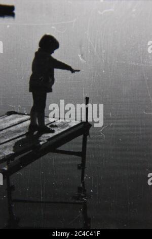 Foto en blanco y negro de un niño parado al final de un muelle mirando las aguas tranquilas. Foto de stock