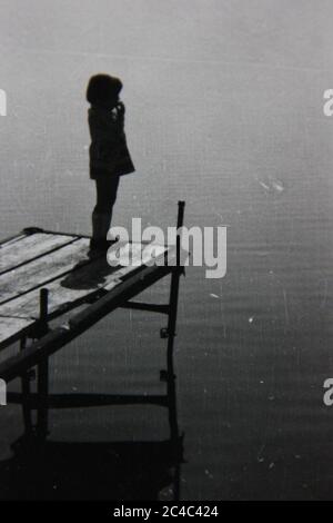 Foto en blanco y negro de un niño parado al final de un muelle mirando las aguas tranquilas. Foto de stock