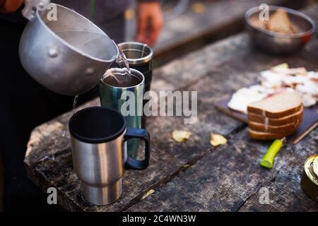 El hombre vierte agua hervida a tazas calientes en una mesa de madera durante el desayuno en el campamento del bosque. Gente en camping picnic en el parque nacional y haciendo té de almuerzo Foto de stock