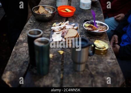 Foto de la mesa de madera de desayuno de excursionistas con pan, tocino, latas, otras comidas y tazas calientes en el campamento de bosque. Gente acampando en el parque nacional y. Foto de stock