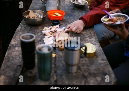 Foto de la mesa de madera de desayuno de excursionistas con pan, tocino, latas, otras comidas y tazas calientes en el campamento de bosque. Gente acampando en el parque nacional y. Foto de stock