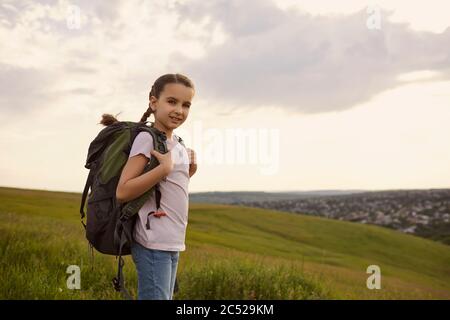 Adorable niña con mochila en la cima de la montaña, espacio de copia. Lindo niño con mochila para hacer senderismo en el campo Foto de stock
