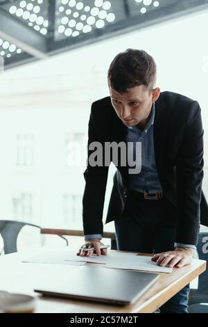 Hombre de negocios joven y seguro que firma documentos en una oficina moderna con ventana en el fondo. Pluma en la mano, papeles en el escritorio de madera Foto de stock