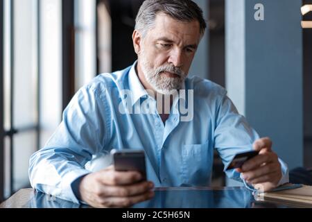 Pago en línea en el smartphone por Mature Man. Guapo líder de negocios maduro sentado en una oficina moderna