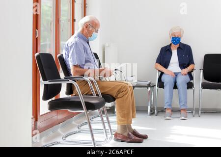 Mujer mayor y hombre sentado con máscaras en una luminosa sala de espera de un hospital o una oficina Foto de stock