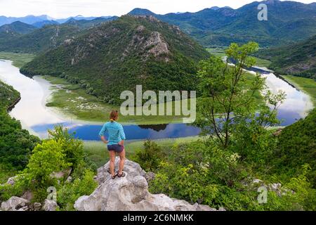 Mujer de pie sobre una roca sobre la curva del río Rijeka Crnojevica, vista desde el mirador de Pavlova strana, cerca de Cetinje Foto de stock