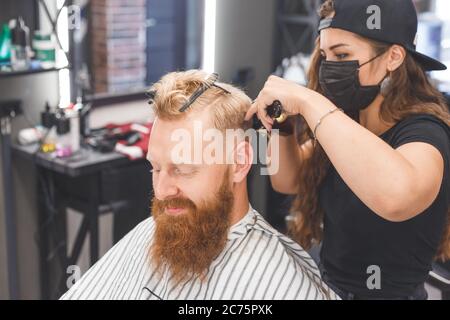 Mujer barbero haciendo corte de pelo al hombre de pelo rojo con barba en la peluquería Foto de stock