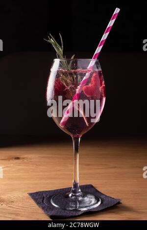 Cóctel frío de color rosa alcohólico con frambuesa con hielo y un tubo en un gran cristal sobre una mesa de madera sobre fondo negro. Foto de stock