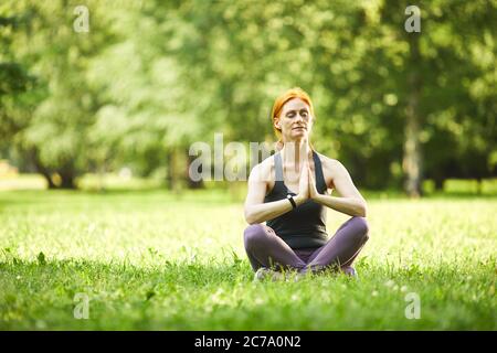 Serena mujer madura de pelirroja sentada sobre hierba y sosteniendo las manos en Namaste mientras meditaba en el parque vacío