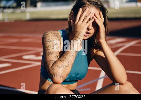 Atractiva chica atleta húmeda en ropa deportiva cuidadosamente cubriendo la cara con las manos mientras descansa después de entrenar en el estadio de la ciudad Foto de stock