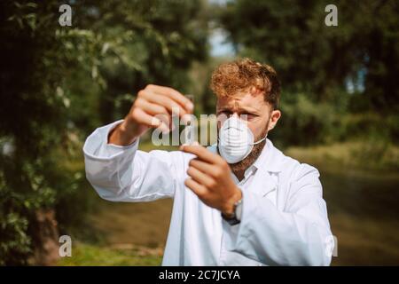 Biólogo científico caucásico masculino e investigador en traje protector con máscara tomando muestras de agua de río contaminado.