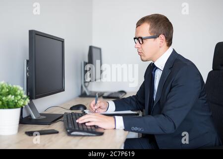 joven hombre de negocios guapo usando la computadora y escribiendo algo en el portapapeles Foto de stock