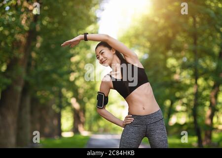 Chica asiática deportiva ejercitando antes de correr en el parque, estirando sus músculos al aire libre