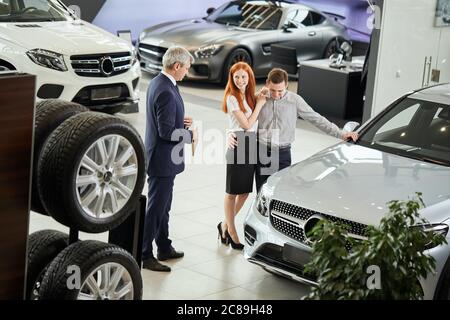 Gerente de ventas profesional maduro ofreciendo pareja joven para tomar drive test en coche elegido al concesionario de coches