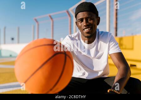 Hombre joven sonriente sosteniendo baloncesto mientras estaba sentado en la cancha durante día soleado