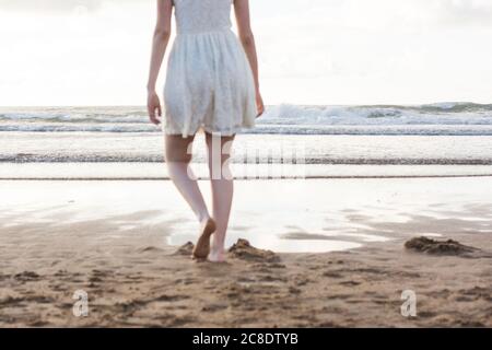Mujer joven con vestido blanco caminando descalzo sobre la arena playa contra el cielo despejado