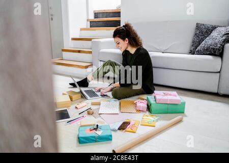 Sonriente mujer joven sentada en el suelo con regalos envueltos usando un portátil