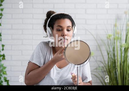 Mujer joven escuchando música a través de auriculares aplicando brillo de labios inicio Foto de stock