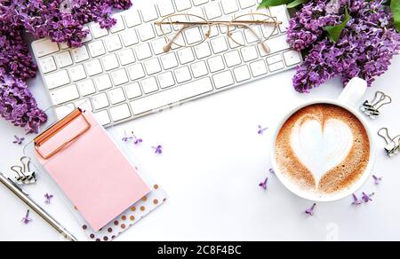 Escritorio de mesa de oficina con vista superior. Espacio de trabajo con teclado, flores lilas y suministros de oficina sobre fondo blanco. Foto de stock
