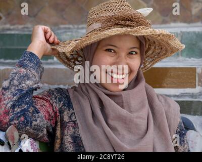 La joven Indonesia lleva un sombrero de paja sobre su hijab tradicional y sonríe para la cámara en el templo Wat Arun de Bangkok. Foto de stock