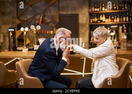 Hombre guapo en el hermoso tuxedo y mujer en blanco blazer sentarse en un restaurante hermoso y caro. Hombre besando la mano y declarando su amor. ima romántica