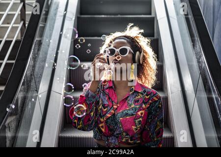 Mujer afroamericana irreconocible en gafas de sol y auriculares inalámbricos soplando burbujas mientras se sienta con labios de boca en escalera mecánica en la parte de atrás iluminada