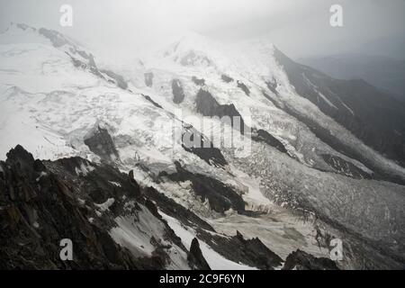 Un glaciar fluye por la ladera de la montaña, con rocas oscuras que sobresalen en primer plano. Nubes de color gris claro se desvían sobre el pico de la montaña.