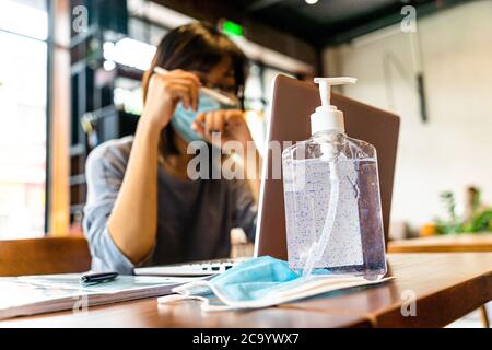 Mujer asiática joven usando computadora portátil trabajando en casa con gel de alcohol en cuarentena para coronavirus usando máscara protectora con distanciamiento social Foto de stock